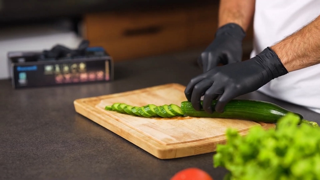 The man is wearing black gloves while cutting a cucumber