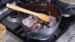 Chopped Onions, Garlic, and Spices Are Being Sautéed in A Pot with A Wooden Spoon on The Stove