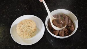 A Plate of Garlic Fried Rice Sits Next to A Bowl of Beef Pares as A Ladle Pours Extra Sauce Over the Beef