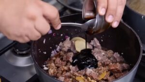 A Person Pours a Dark Sauce Into a Pot of Simmering Beef, Ginger, and Onions While Stirring with A Spoon